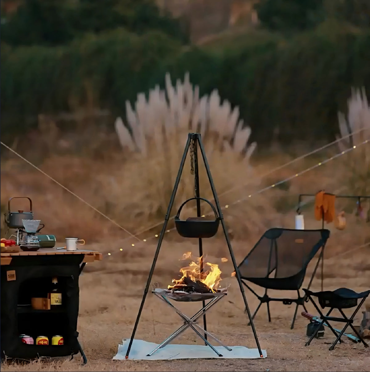 The fire pit burning at a campsite beside a table and chairs, under a cooking tripod with a hanging pot (tripod and pot not included)